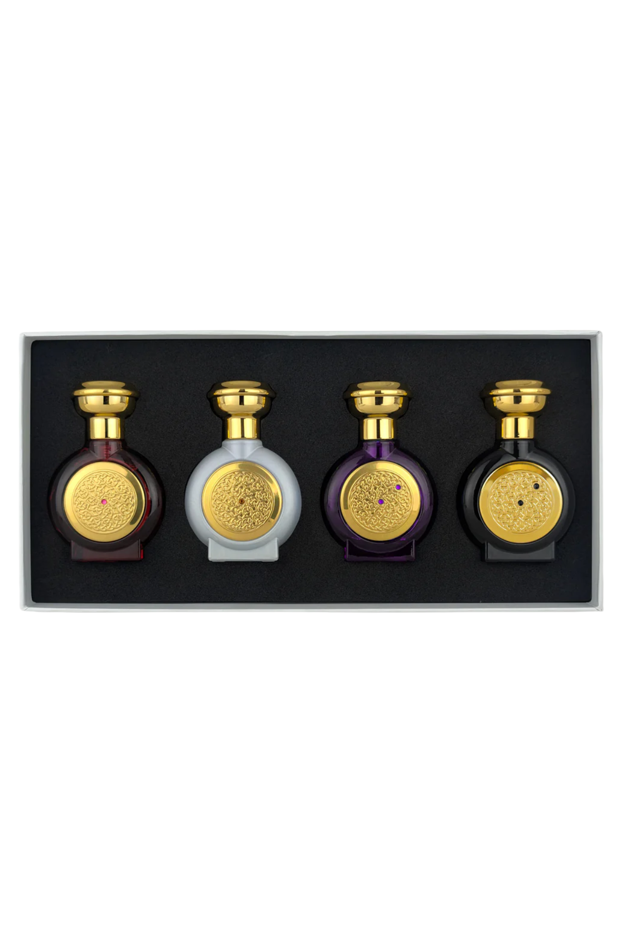 A boxed set of four decorative bottles arranged in a black tray against a white background. Three bottles are dark purple with gold caps and round gold medallions, and one bottle is pale gray with a gold cap and medallion.
