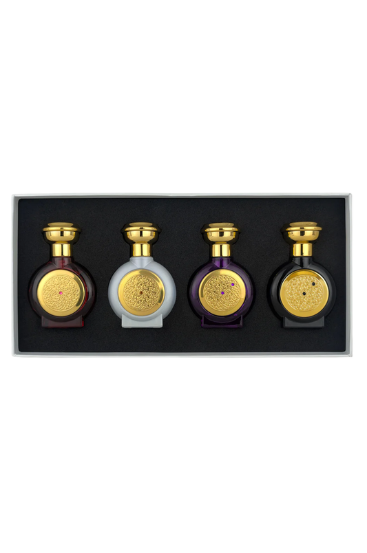 A boxed set of four decorative bottles arranged in a black tray against a white background. Three bottles are dark purple with gold caps and round gold medallions, and one bottle is pale gray with a gold cap and medallion.